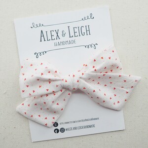 Peut inclure: Un n&oelig;ud en tissu blanc avec un motif de c&oelig;urs rouges. Le n&oelig;ud est nou&eacute; avec un ruban blanc et porte une petite &eacute;tiquette blanche avec le texte "Alex & Leigh Handmade" et des informations sur les r&eacute;seaux sociaux.