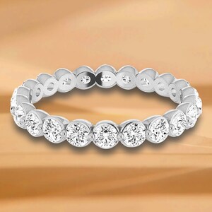 Könnte beinhalten: Ein silberner Eternity-Ring mit einer durchgehenden Reihe runder, brillantgeschliffener Diamanten. Die Diamanten sind eng aneinander gesetzt und bilden ein funkelndes Band. Der Ring wird vor einem warmen, goldbraunen Hintergrund präsentiert.
