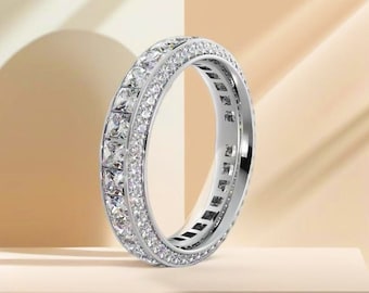 May include: A silver ring with two rows of square-cut, clear gemstones. The ring is encrusted with smaller, round gemstones between the rows. The ring is set against a neutral background, highlighting its sparkle and design.