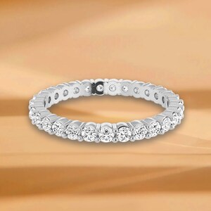 Könnte beinhalten: Ein silberner Eternity-Ring mit einer durchgehenden Reihe runder, klarer Edelsteine. Der Ring ist vor einem warmen, goldbraunen Hintergrund platziert, der das Funkeln der Steine hervorhebt. Das Design des Rings ist schlicht und elegant.