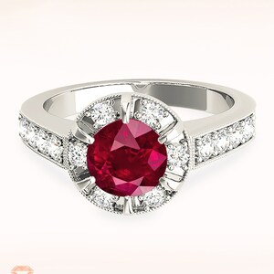 Op de afbeelding: Een zilveren ring met een grote rode robijn-edelsteen omringd door een halo van kleinere diamanten. De band is ook bezet met kleinere diamanten.
