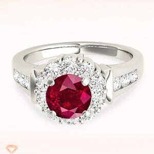 Op de afbeelding: Een zilveren ring met een grote rode robijn, omringd door een halo van kleinere diamanten. De band is bezet met extra diamanten.