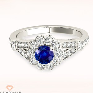 Könnte beinhalten: Ein silberner Ring mit einem blauen Saphir als Mittelstück, umgeben von einem Heiligenschein aus kleineren Diamanten. Der Ring hat ein zartes Blumenmuster und ist mit zusätzlichen Diamanten auf der Ringschiene besetzt.