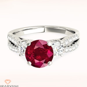 May include: A silver ring featuring a large red ruby gemstone, flanked by two smaller diamonds. The ring has a split shank with pave diamonds on each side.