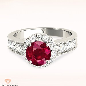 Op de afbeelding: Een zilveren ring met een grote rode robijn-edelsteen omringd door een halo van kleinere diamanten. De band is ook bezet met diamanten.