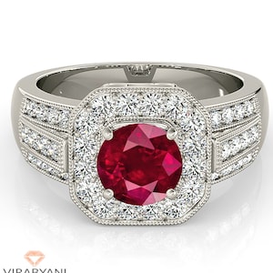 以下が含まれることがあります： 中央に大きな丸い深紅色の宝石をあしらったシルバーリング。周囲には小さな透明な宝石が散りばめられています。リングは正方形のハローデザインで、バンドにも宝石が施されています。「VIRABYANI FINE JEWELRY」のブランド名が下部にあります。