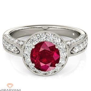 Op de afbeelding: Een zilveren ring met een grote rode robijn omringd door een halo van kleinere diamanten. De band is versierd met extra diamanten.