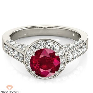 Op de afbeelding: Een zilveren ring met een grote rode robijn-edelsteen omringd door een halo van kleinere diamanten. De band is ook bezet met diamanten.