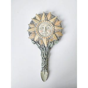 May include: A hand mirror featuring a sun face at the center with radiating triangular rays. The handle is sculpted with leaf and vine details in a muted, weathered beige and grey finish. The overall design is circular with a long stem.