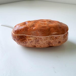 May include: A ceramic lidded container shaped like a russet potato with a textured, mottled brown and tan glazed surface. The oval-shaped vessel features a removable lid and a small white ceramic spoon protruding from the side of the base.
