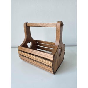 May include: A medium-brown wooden caddy with a slatted design and a central handle. The two side panels feature a cutout heart shape. The structure is composed of horizontal wooden slats forming a rectangular base and open sides for storage.