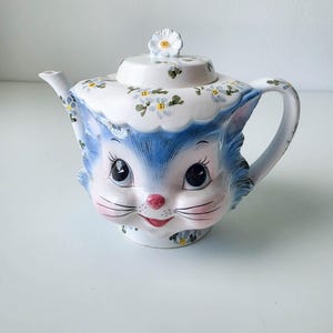 May include: A ceramic teapot shaped like a cat head with blue fur, large dark eyes, a pink nose, and whiskers. The lid features a white flower handle, and the entire piece is decorated with small blue and yellow floral patterns on a white base.