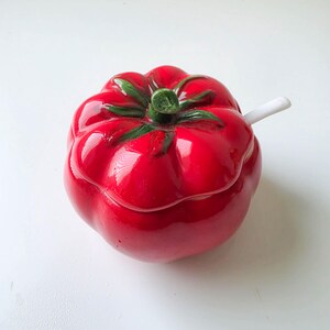 May include: A glossy red ceramic lidded jar shaped like a tomato with a green stem handle on the lid. A small white ceramic spoon is tucked into the side of the jar, resting between the lid and the base, providing a functional serving utensil.