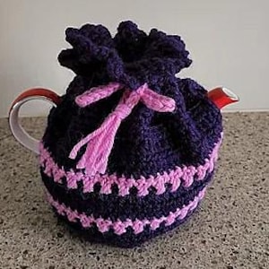 May include: A dark purple crocheted tea cosy with a light pink trim and drawstring closure. The cosy covers a white teapot with a red handle and spout.