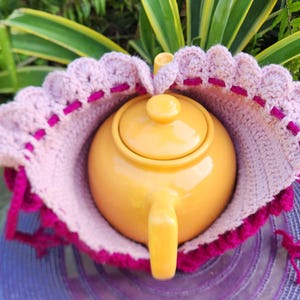 May include: A pink and purple crocheted teapot cozy with a yellow teapot inside. The cozy has a scalloped edge and is made with a textured stitch.
