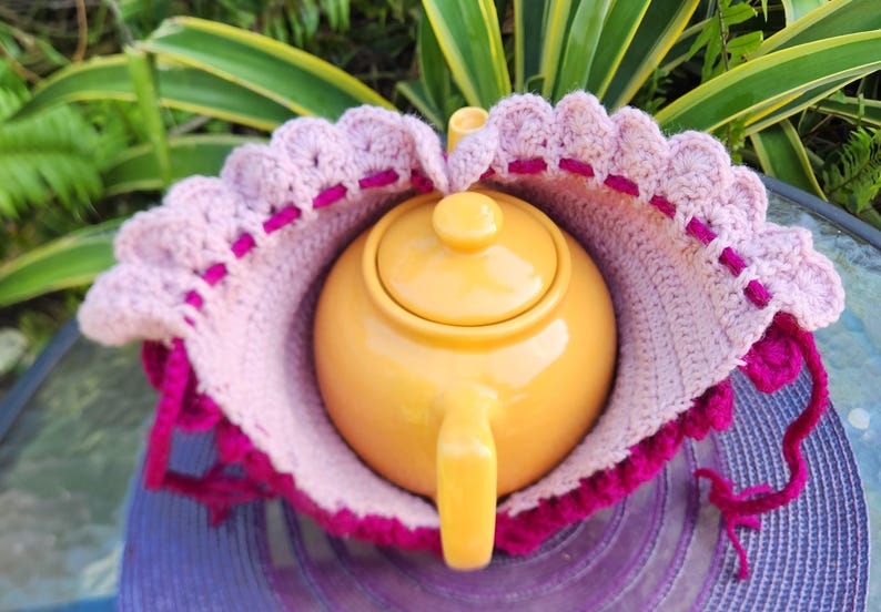 May include: A pink and purple crocheted teapot cozy with a yellow teapot inside. The cozy has a scalloped edge and is made with a textured stitch.