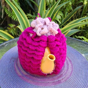 May include: A pink crocheted teapot cozy with a flower design on top. The cozy is covering a yellow teapot.