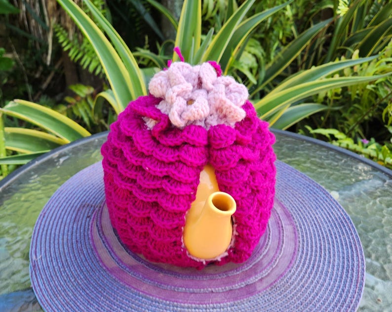 May include: A pink crocheted teapot cozy with a flower design on top. The cozy is covering a yellow teapot.