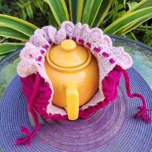 May include: A yellow teapot with a pink and magenta crocheted tea cozy. The cozy has a ruffled edge and a long pink string.