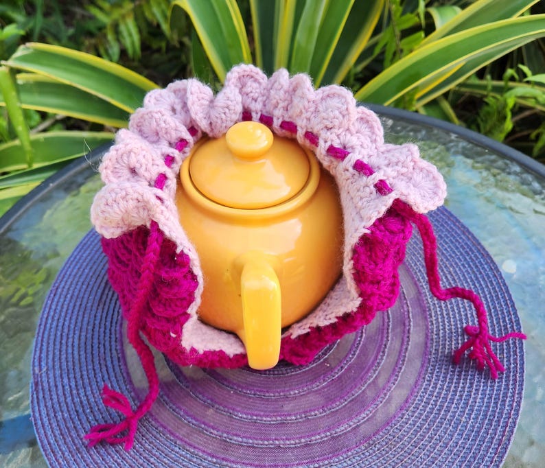 May include: A yellow teapot with a pink and magenta crocheted tea cozy. The cozy has a ruffled edge and a long pink string.