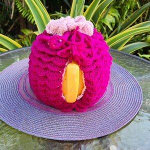 May include: A crocheted pink and purple teapot cozy with a yellow handle. The cozy is sitting on a purple and white straw hat.