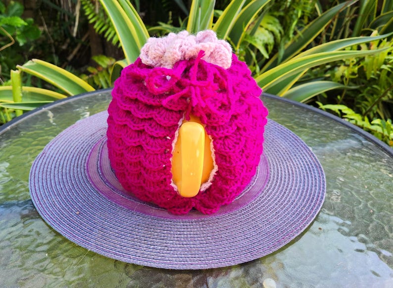 May include: A crocheted pink and purple teapot cozy with a yellow handle. The cozy is sitting on a purple and white straw hat.