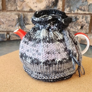 May include: A gray and white knitted teapot cozy with a black trim. The cozy is on a red teapot with a white handle.