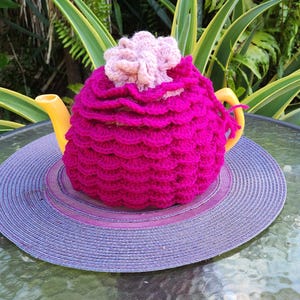 May include: A pink and purple crocheted teapot cozy with a flower on top. The cozy is on a yellow teapot and a purple woven coaster.