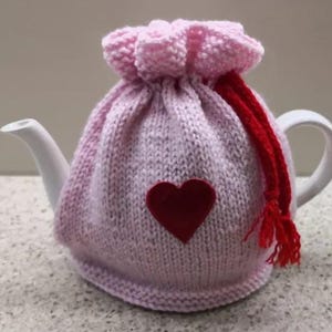 May include: Pink knitted teapot cozy with a red heart design and a red drawstring.