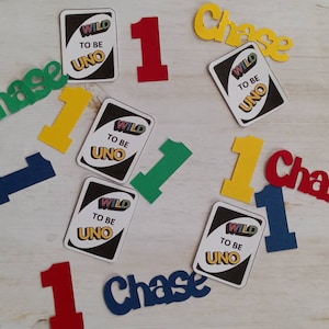 May include: Scattered on a light wood surface are colorful cutouts for a birthday celebration. The cutouts include the name "Chase" in green, yellow, and red, along with the number "1" in red, yellow, green, and blue, and "Wild to be Uno" cards.