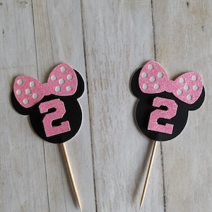 May include: Two black and pink Minnie Mouse-themed cupcake toppers. Each topper features a black silhouette of Minnie's head, a pink bow with white polka dots, and the number "2" in pink glitter. The toppers are on wooden sticks.