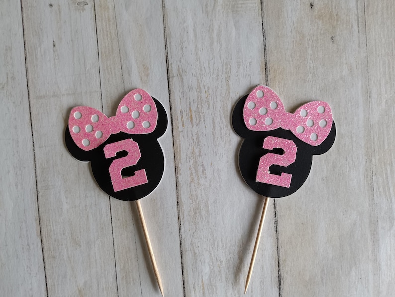 May include: Two black and pink Minnie Mouse-themed cupcake toppers. Each topper features a black silhouette of Minnie's head, a pink bow with white polka dots, and the number "2" in pink glitter. The toppers are on wooden sticks.