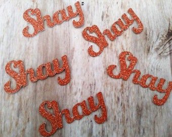 May include: Several orange glitter-covered cutouts spelling the name "Shay" are scattered on a light brown wooden surface. The letters are in a cursive font, and the glitter gives them a sparkling appearance. The cutouts are likely used for crafts or decorations.