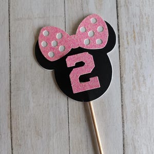 May include: A black and pink Minnie Mouse-themed number two cake topper. The topper features a pink bow with white polka dots and a pink number 2. The topper is attached to a wooden stick.
