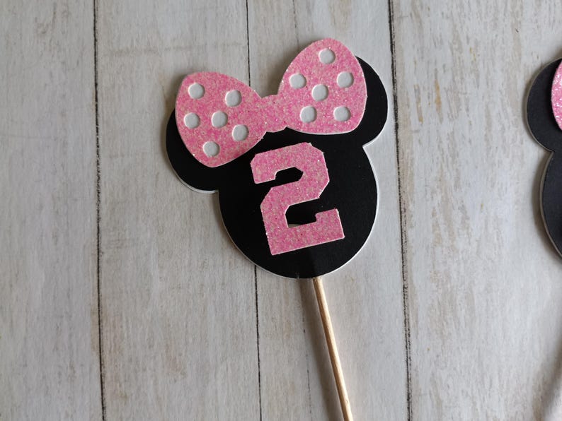 May include: A black and pink Minnie Mouse-themed number two cake topper. The topper features a pink bow with white polka dots and a pink number 2. The topper is attached to a wooden stick.
