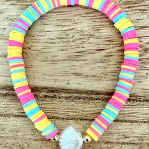 May include: A stretch bracelet made of small, flat, circular polymer clay beads in alternating bright yellow, hot pink, and light blue colors. The bracelet features a single white, iridescent seashell-shaped charm centered between two small gold-toned beads.