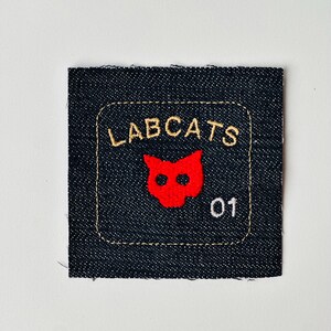Può includere: Toppa quadrata in denim con testo ricamato e una faccia di gatto rossa. Il testo "LABCATS" è in filo d'oro sopra la faccia del gatto, con il numero "01" sotto. La toppa ha un bordo cucito.