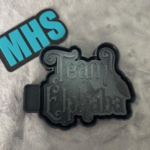 Puede incluir: Un cortador de galletas de plástico negro impreso en 3D con el texto "Team Alpha" en una fuente decorativa.