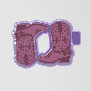 Könnte beinhalten: Ein lila cowboy-stiefelförmiges Objekt mit einem Fleur-de-Lis-Design und Quastenakzenten. Der Stiefel hat ein dekoratives Muster aus Punkten und ist in einer helleren lila Farbe umrandet. Das Objekt befindet sich auf weißem Hintergrund.