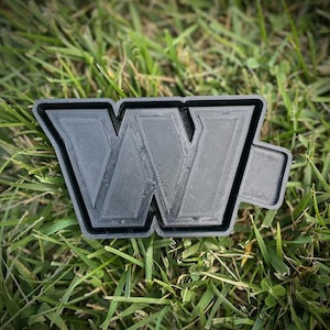 May include: A black plastic 3D printed letter W. The letter is sitting on a bed of green grass.