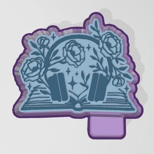 May include: A layered, die-cut bookmark featuring a light blue design of headphones, roses, and an open book. The bookmark has a purple border and a rectangular tab at the bottom. The design includes stars and floral elements.