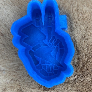 May include: A blue mould in the shape of a hand making a peace sign, with a lightning bolt design. The mould is made of a solid, smooth material and is set against a brown, furry background.