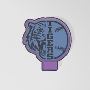 May include: A purple and blue baseball-themed design featuring a tiger's face on the left and a baseball on the right. The word "TIGERS" is vertically centered. The design has a purple border and a purple base.