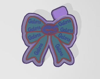 May include: A purple and teal bow-shaped sticker with the word "Gators" repeated multiple times. The bow has a red outline and a purple border. The center features a Gator logo.