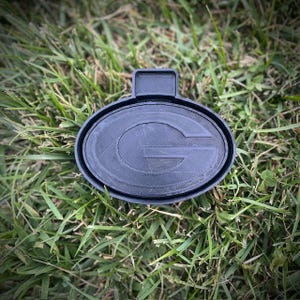 Puede incluir: Un adorno de plástico negro con forma de balón de fútbol con una letra "G" en relieve en el centro. El adorno está sobre un lecho de hierba verde.