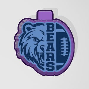 Puede incluir: Un adorno azul y morado con la cabeza de un oso y un balón de fútbol. La palabra "BEARS" se muestra verticalmente. El oso es azul claro, con un contorno azul oscuro. El balón de fútbol es azul oscuro con cordones blancos.