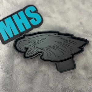 May include: A black plastic cookie cutter shaped like an eagle's head. The eagle's beak is open and the wings are spread out. The cookie cutter is on a grey surface.