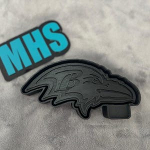 May include: Black plastic cookie cutter shaped like a raven with the letter "B" inside the wing. The cookie cutter is on a gray surface.
