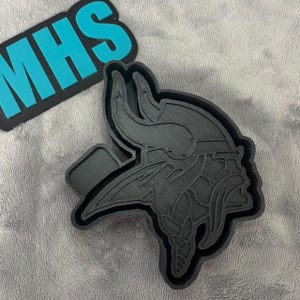 May include: A black 3D printed cookie cutter shaped like a Viking helmet. The helmet has a detailed design with horns and a face mask.