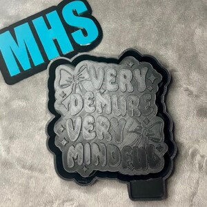 Puede incluir: Un letrero negro impreso en 3D con el texto "Very Demure Very Mindful" en una fuente estilizada. El letrero tiene dos pequeños lazos a cada lado del texto.